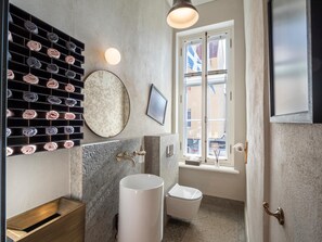 A modern bathroom with a marble sink, a toilet, a towel rack with neatly rolled towels, a round mirror, and a window with a view of buildings.