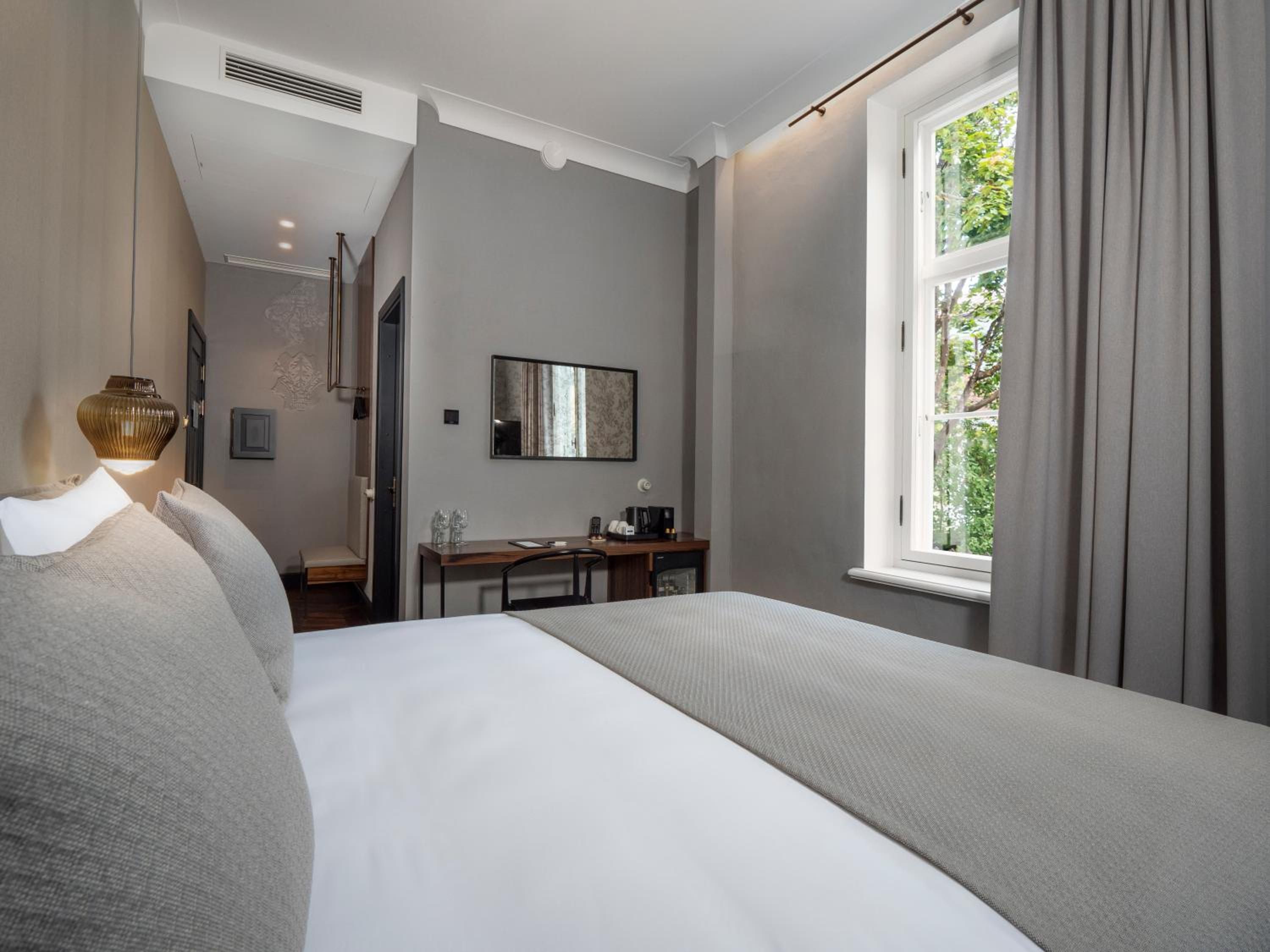 A hotel room with a bed, a desk, a chair, a window with curtains, and a wall-mounted mirror.