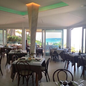 A restaurant with multiple tables and chairs, large windows, and a view of the ocean. - Hotel Boschetto (Follonica)