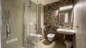 A bathroom with a glass-enclosed shower, a toilet, a bidet, and a sink with a stone backsplash. - Hotel Boschetto (Follonica)