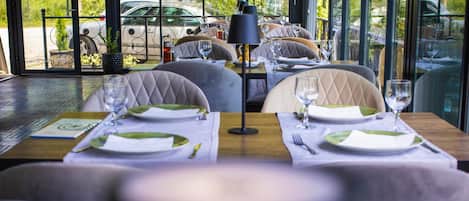 Un restaurant avec des tables dressées, une vue sur les voitures garées à l’extérieur et un panneau indiquant les heures d’ouverture du parking.