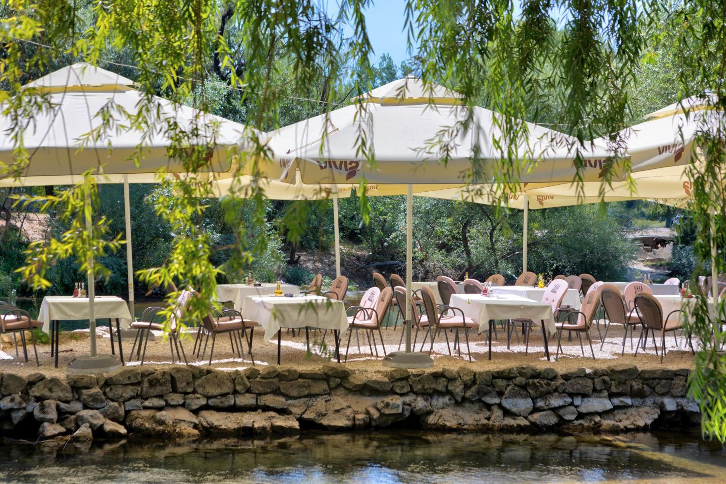 Un área de comedor al aire libre con mesas y sillas bajo sombrillas, un muro de piedra y una masa de agua.