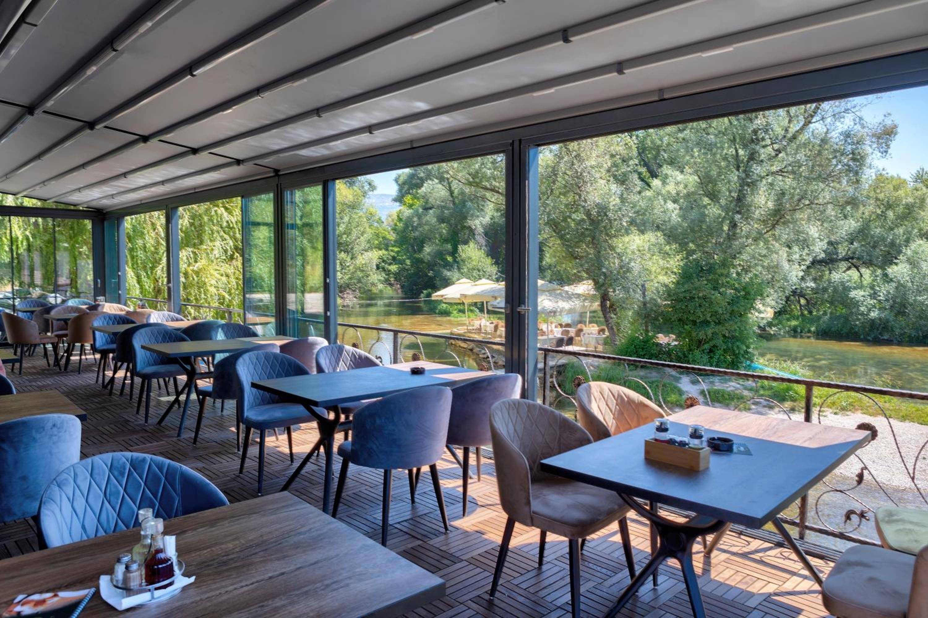 Un espacio de comedor al aire libre con mesas y sillas, con vistas a un río y follaje.