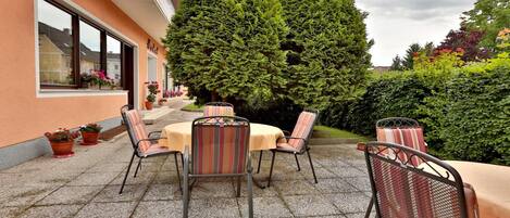 An outdoor patio with tables and chairs, surrounded by potted plants and a large tree.