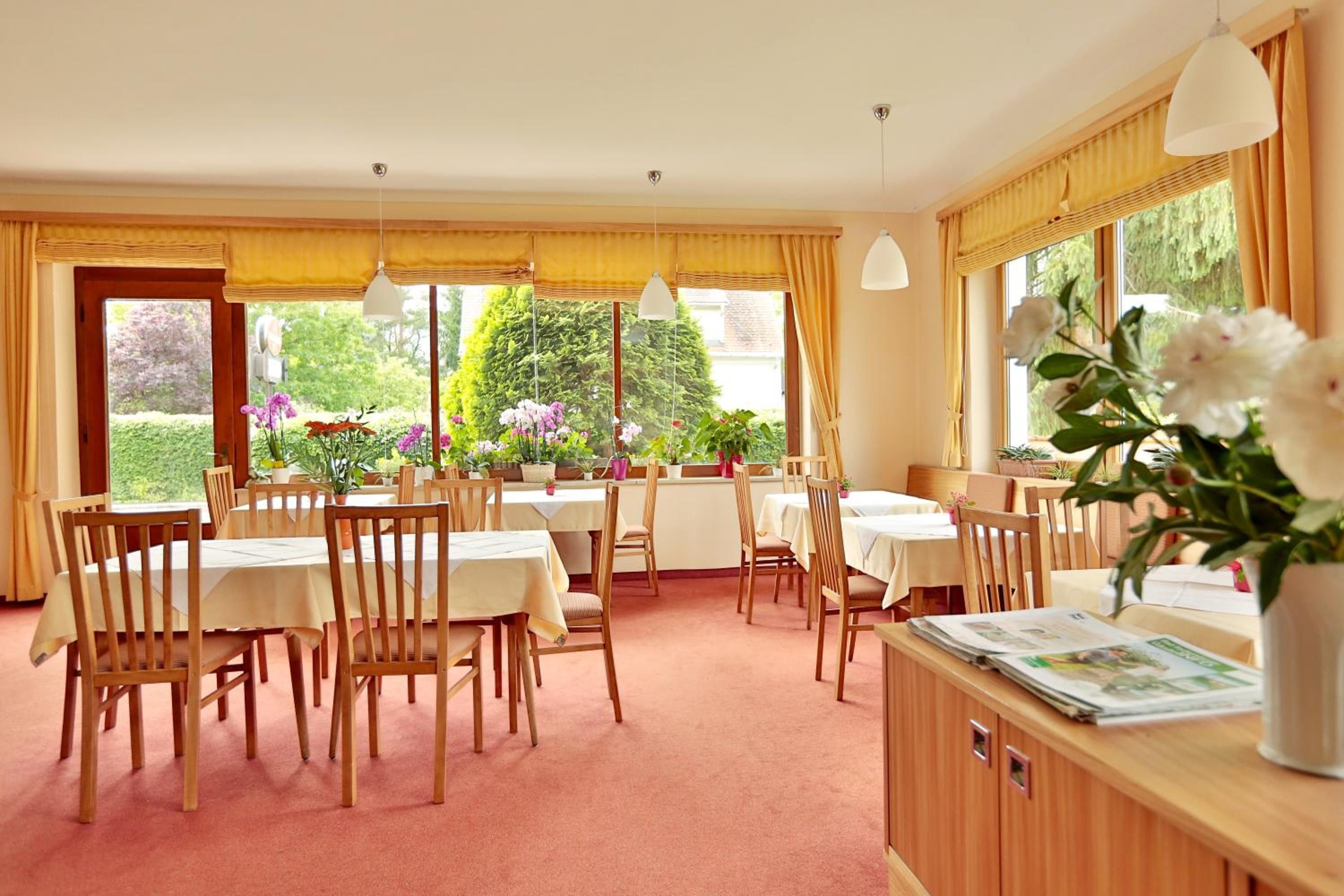 Un espace repas avec des tables et des chaises en bois, de grandes fenêtres avec des rideaux et une vue sur la verdure à l’extérieur.