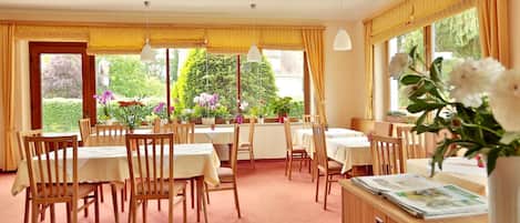 Un espace repas avec des tables et des chaises en bois, de grandes fenêtres avec des rideaux et une vue sur la verdure à l’extérieur.