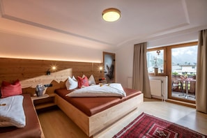 Un dormitorio con cabecera de madera, una cama grande con múltiples almohadas, una mesita de noche, una pequeña mesa con un florero y vistas al exterior a través de una ventana.