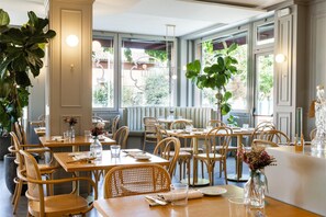 A restaurant interior with wooden tables and chairs, large windows, and indoor plants. - Hotel Emi (Calderara di Reno)