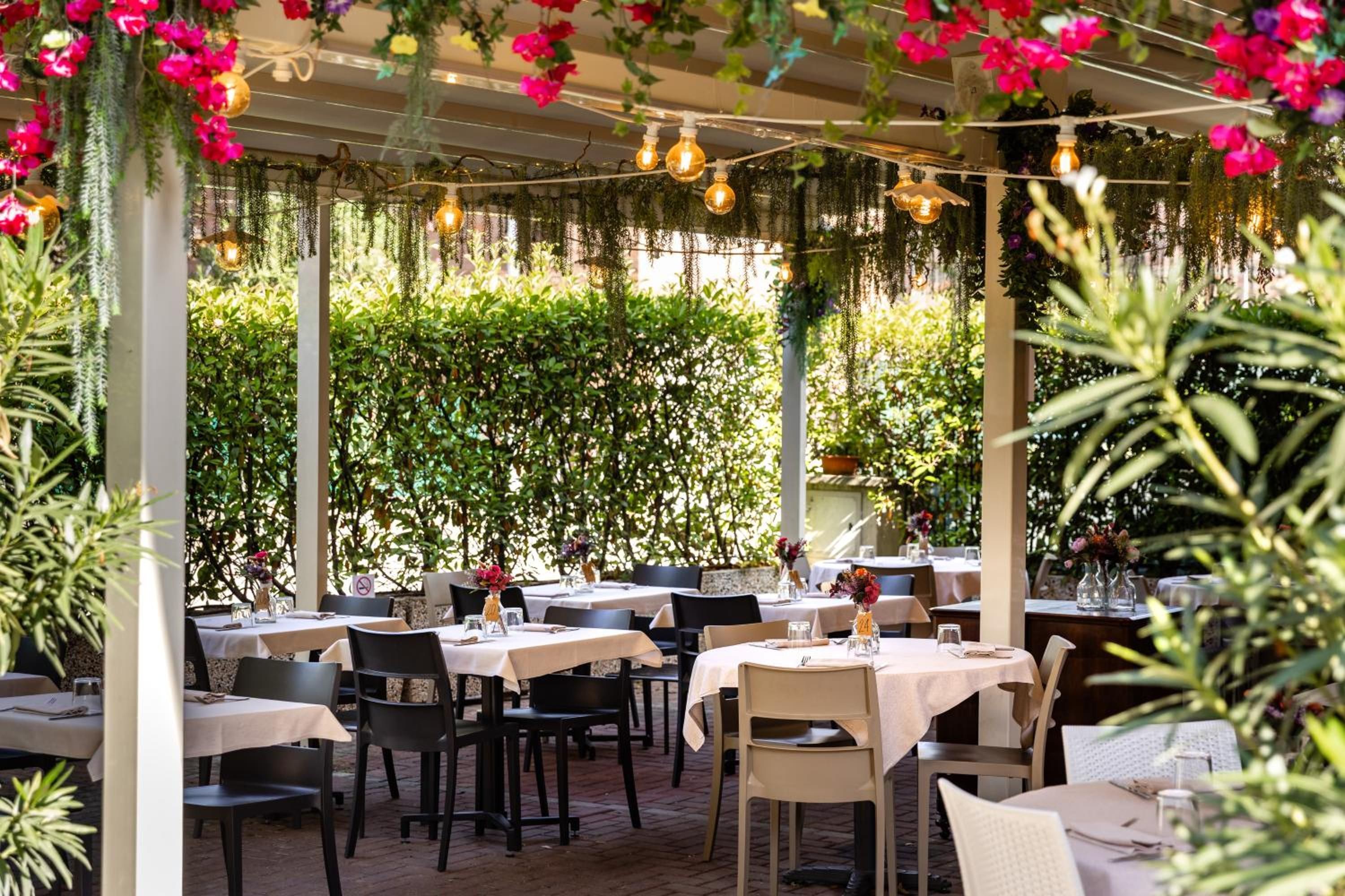 Un espacio de comedor al aire libre con mesas y sillas, rodeado de follaje y plantas colgantes.