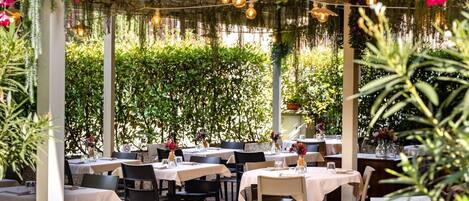 An outdoor dining area with tables and chairs, surrounded by greenery and hanging plants.