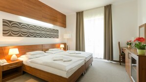 A hotel room with a double bed, two bedside tables with lamps, a large abstract wall art piece, a window with curtains, and a small desk with a chair and a potted plant. - Hotel Kelep (Tokaj)
