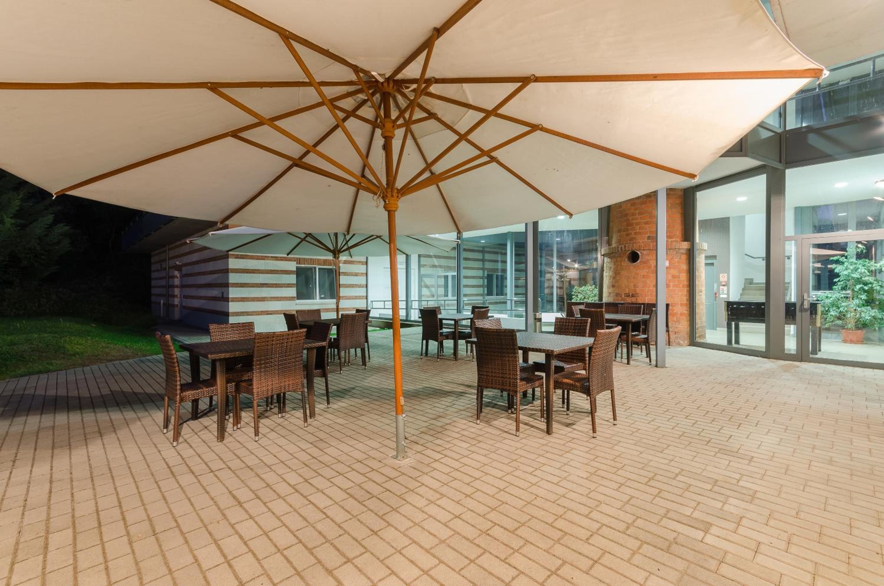An outdoor patio with tables and chairs under a large umbrella, surrounded by brick flooring and glass doors.