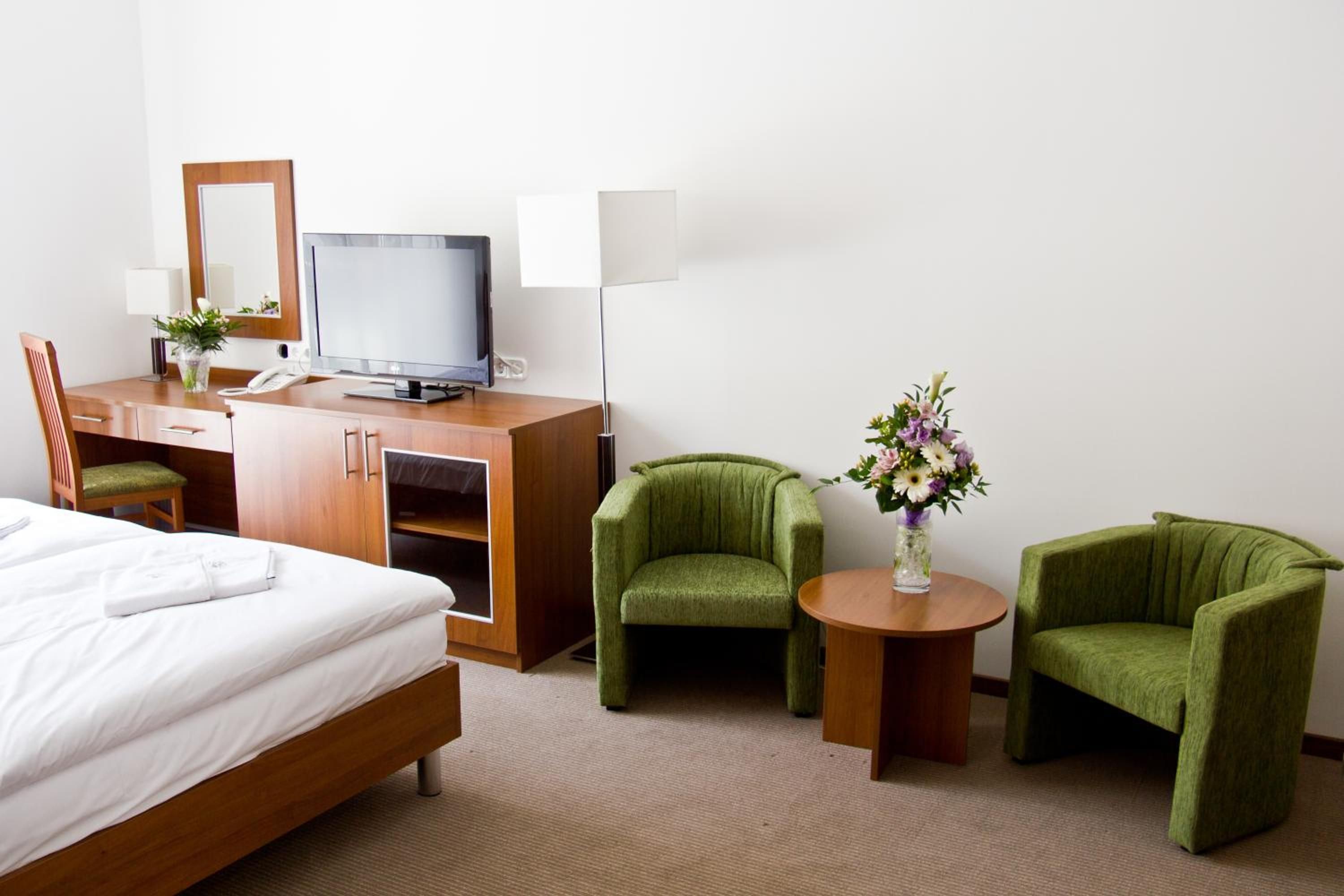 A hotel room with a bed, a desk with a computer, two green armchairs, a small table with a vase of flowers, and a mirror.