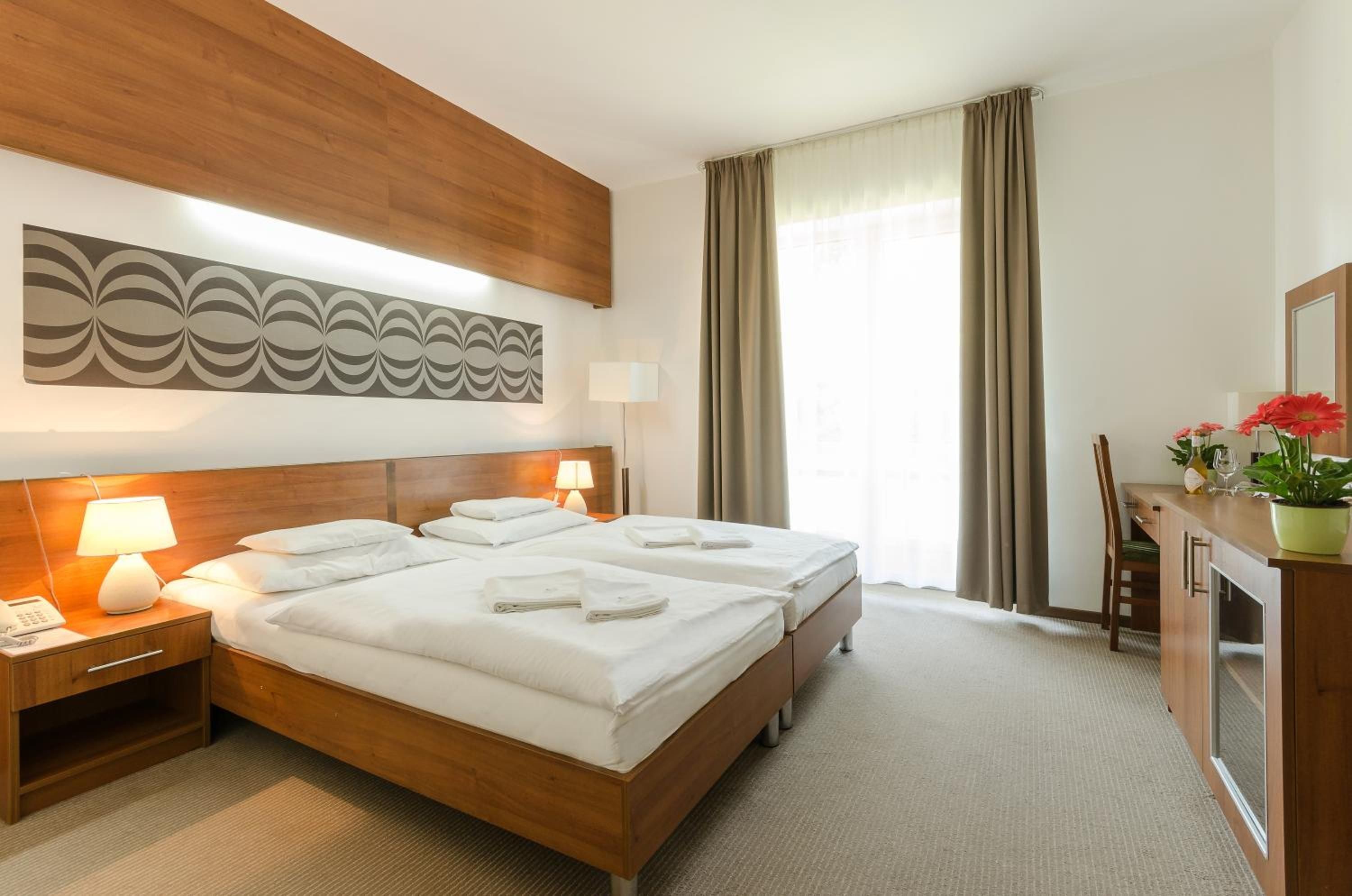 A hotel room with a double bed, two bedside tables with lamps, a large abstract wall art piece, a window with curtains, and a small desk with a chair and a potted plant.