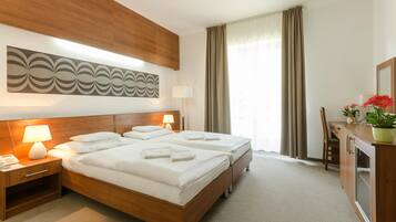 A hotel room with a double bed, two bedside tables with lamps, a large abstract wall art piece, a window with curtains, and a small desk with a chair and a potted plant.
