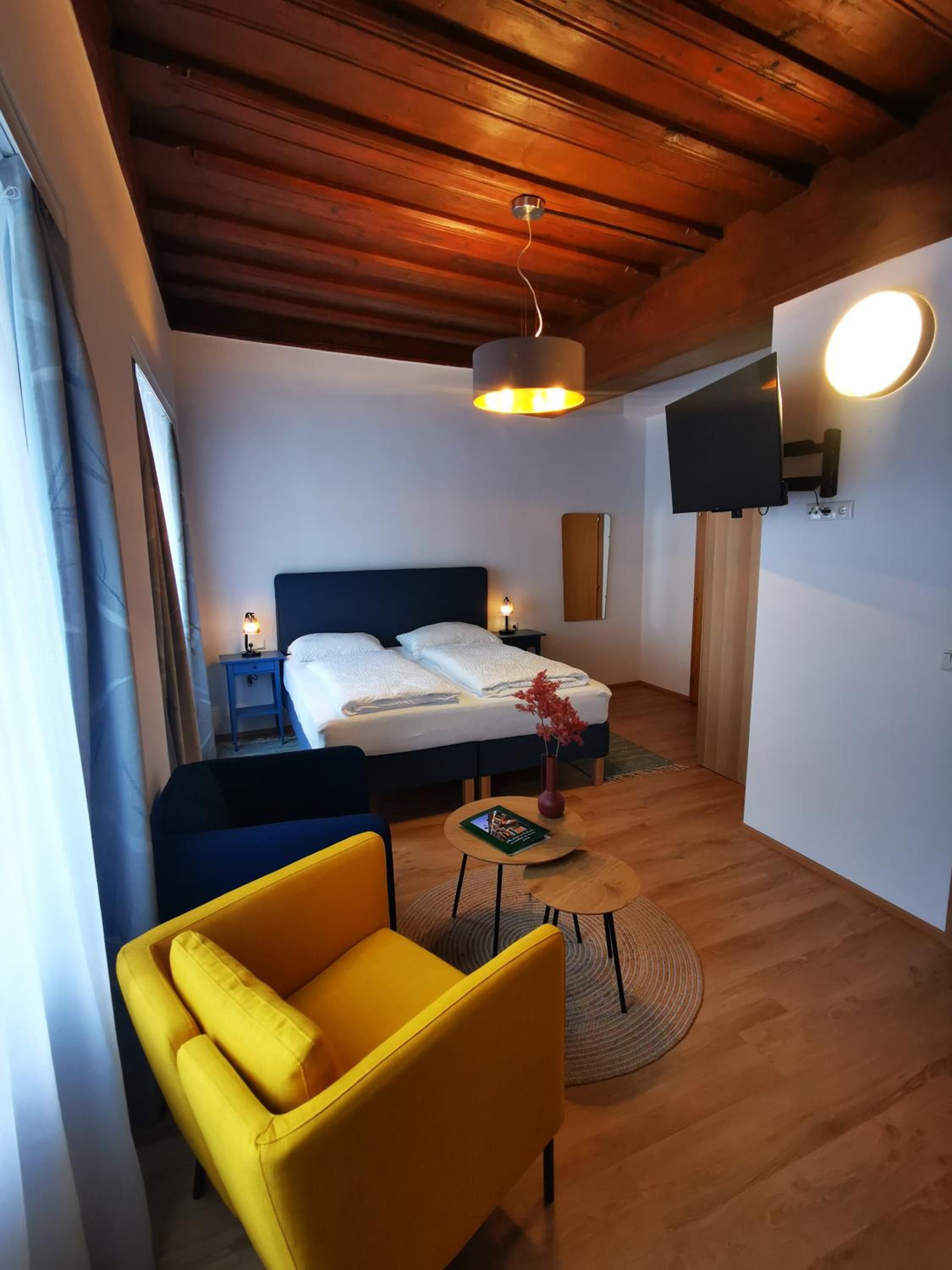 A hotel room with a bed, a yellow armchair, a small table, a TV mounted on the wall, and a wooden ceiling.