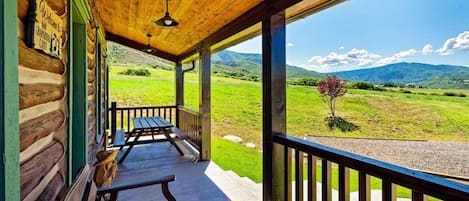 A wooden cabin with a porch, a bench, and a view of a green landscape with mountains.