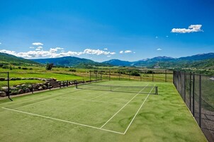 Una cancha de tenis con una red, rodeada por una cerca, y un paisaje montañoso al fondo.