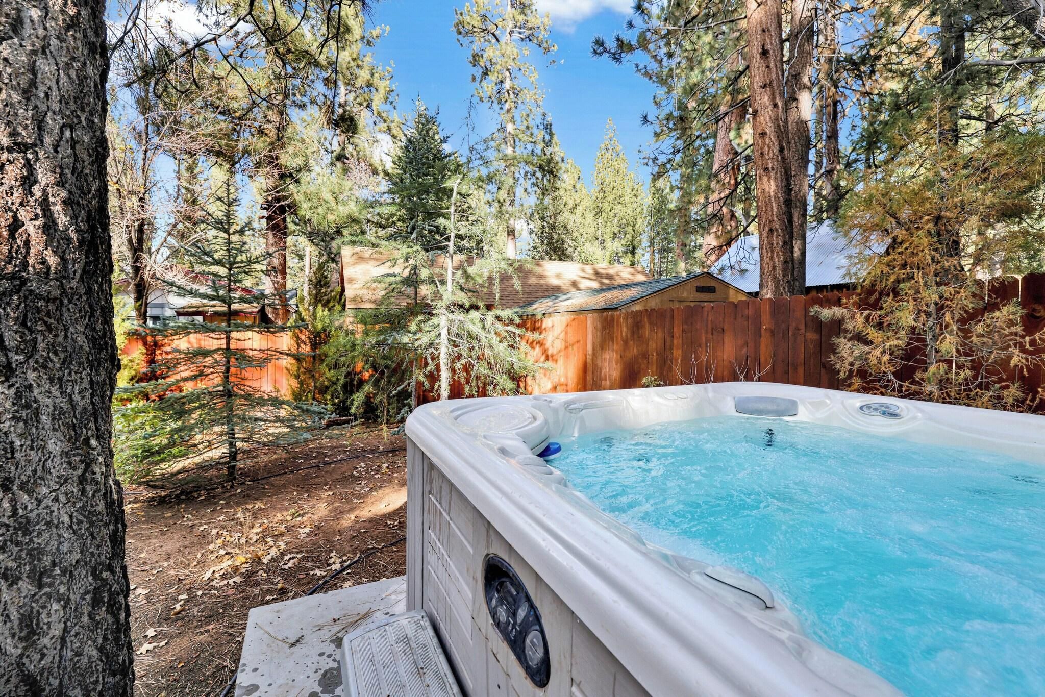 A hot tub in a forested backyard with a wooden fence and a shed in the background.