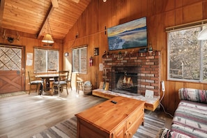 A cozy cabin interior with a fireplace, wooden walls, a dining area, and large windows. - Joebellarue Home (Big Bear Lake)