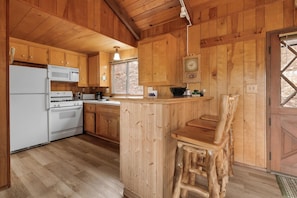 A wooden cabin kitchen with a central island, wooden cabinets, a microwave, stove, and a clock on the wall. - Joebellarue Home (Big Bear Lake)