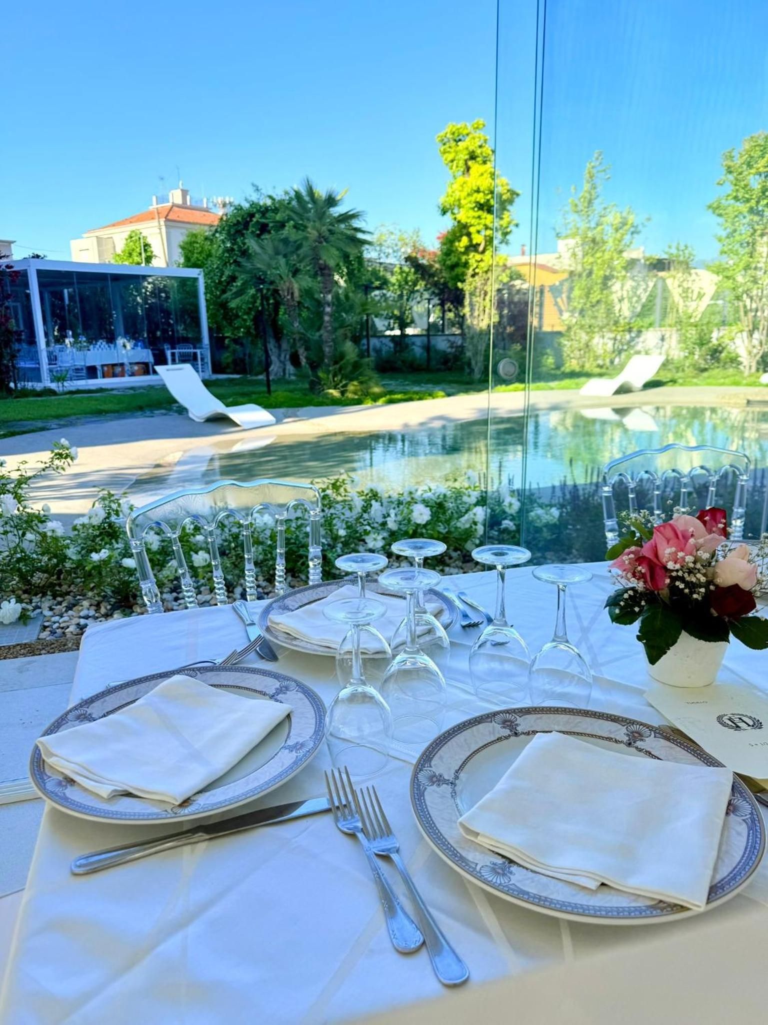 An outdoor dining setup with a view of a pool and greenery, including a table with plates, glasses, and cutlery.