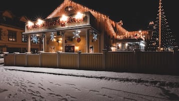 Un hôtel dont le sol est recouvert de neige, avec un balcon illuminé et un sapin de Noël en arrière-plan.