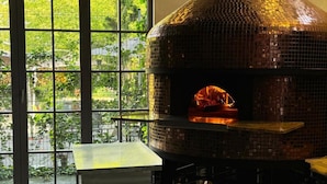 Een keuken met een grote, betegelde pizzaoven, een geruit vloertje en een raam met uitzicht op groen.