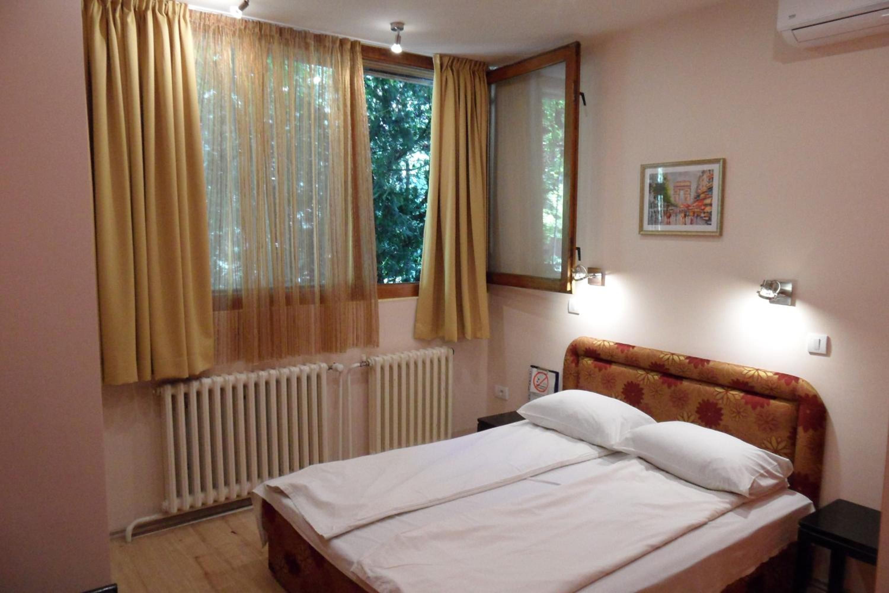 A bedroom with a bed, a window with curtains, a radiator, a wall-mounted light, and a framed picture.