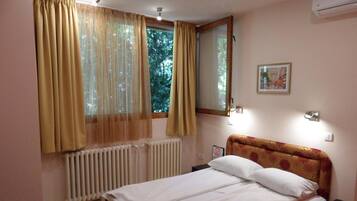 A bedroom with a bed, a window with curtains, a radiator, a wall-mounted light, and a framed picture.