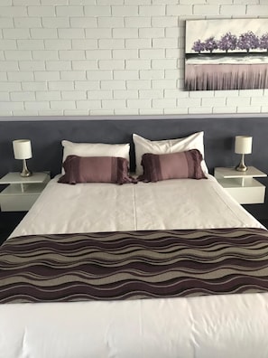 A hotel room with a white brick wall, a bed with white and brown bedding, two bedside tables with lamps, and a painting above the bed.