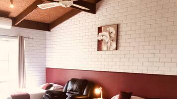 A hotel room with a large bed, a chair, a bedside table with a lamp, a ceiling fan, and a brick-patterned wall.