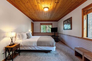 Un dormitorio con techo de madera, una cama, una mesita de noche con lámpara, un televisor y un ventanal con vista a árboles.