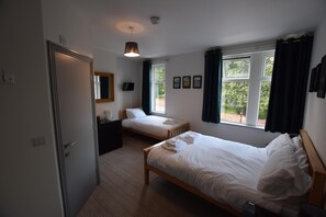 A hotel room with two single beds, a bedside table, a mirror, a TV, and a window with curtains. - The Crow Inn (Sheffield)