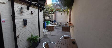 Un patio estrecho con una mesa y sillas de madera, un muro de ladrillo y una escalera que sube a un nivel superior.