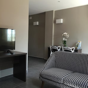 A modern living room with a flat-screen TV, a black and white patterned sofa, and a dining area with white chairs. - Le Brin D Olivier (Bourg-Saint-Andeol)