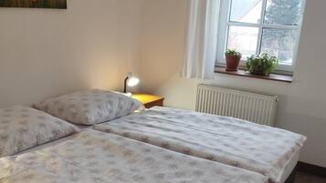 A bedroom with two beds, a bedside table with a lamp, a window with curtains, and a radiator.