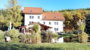 A two-story building with a white facade and red-tiled roof, situated beside a calm lake with surrounding greenery.