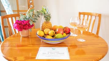 Una mesa de madera para comer con un frutero, una piña, un jarrón de flores y dos copas de vino. Hay una tarjeta de bienvenida sobre la mesa.