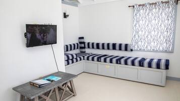 Een kamer met een aan de muur bevestigde televisie, een bank met gestreepte kussens, een klein tafeltje met een boek en een kamerbreed gordijn.