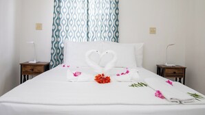 A hotel room with a bed, two bedside tables, a decorative swan towel, and a patterned curtain. - Neem View Apartments (Falmouth Harbour)