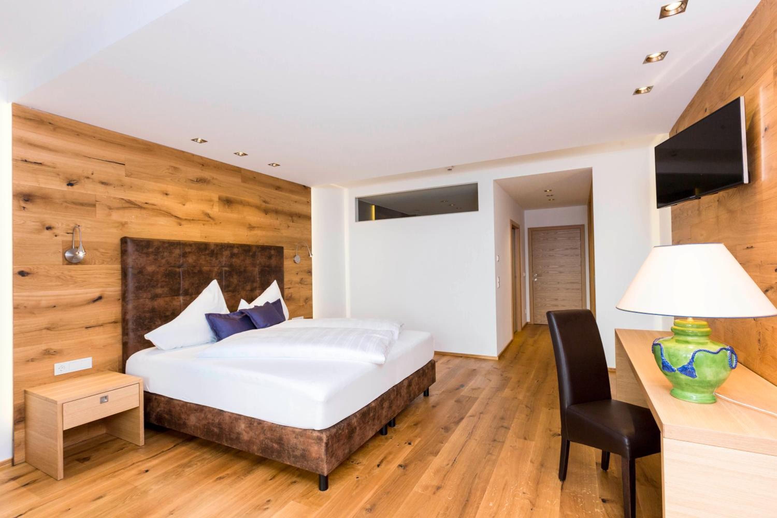 A modern hotel room with a wooden headboard, a bed with white linens, a desk with a lamp, and a television mounted on the wall.