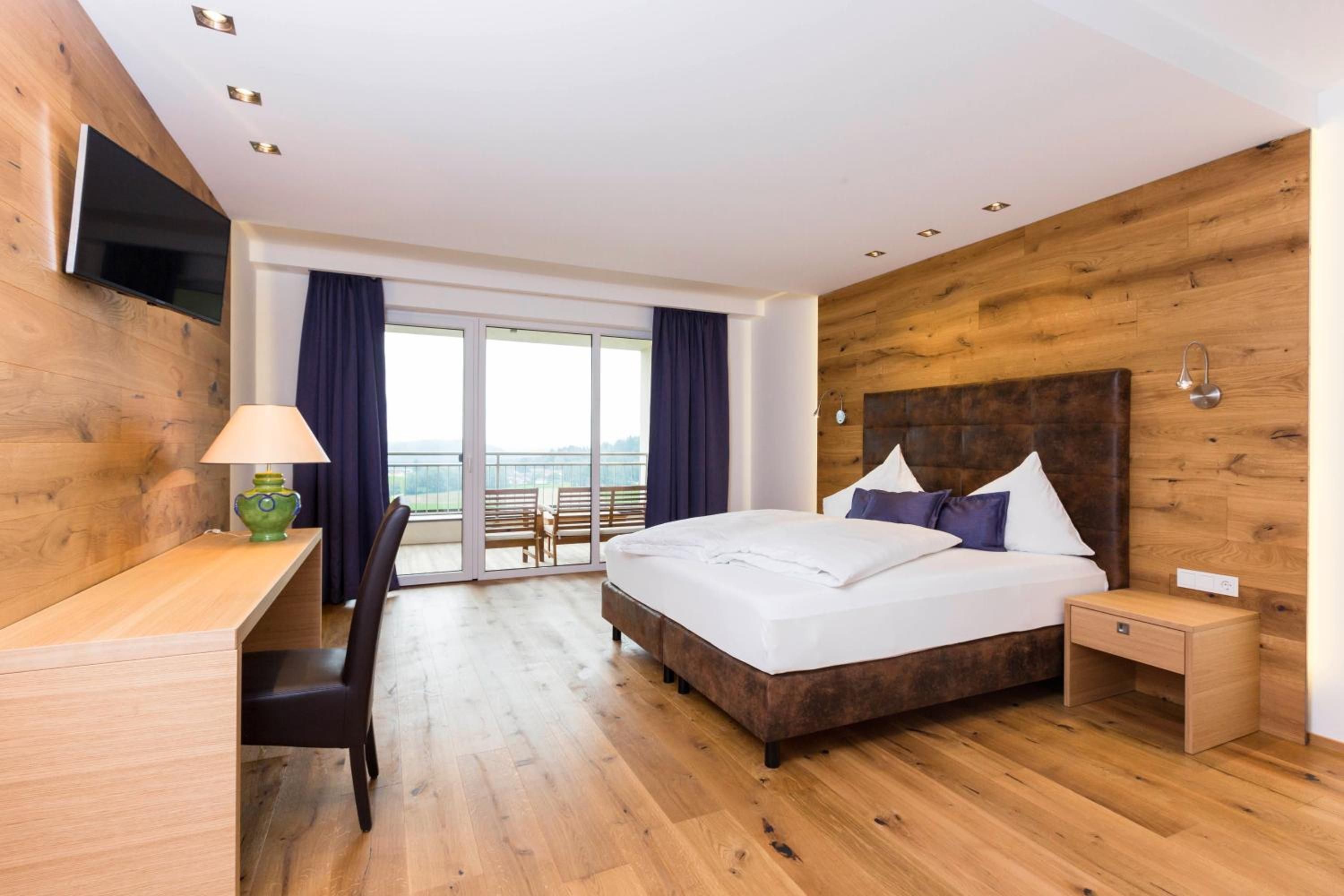 A hotel room with a wooden floor, a bed with white linens, a desk with a lamp, and a TV mounted on the wall.