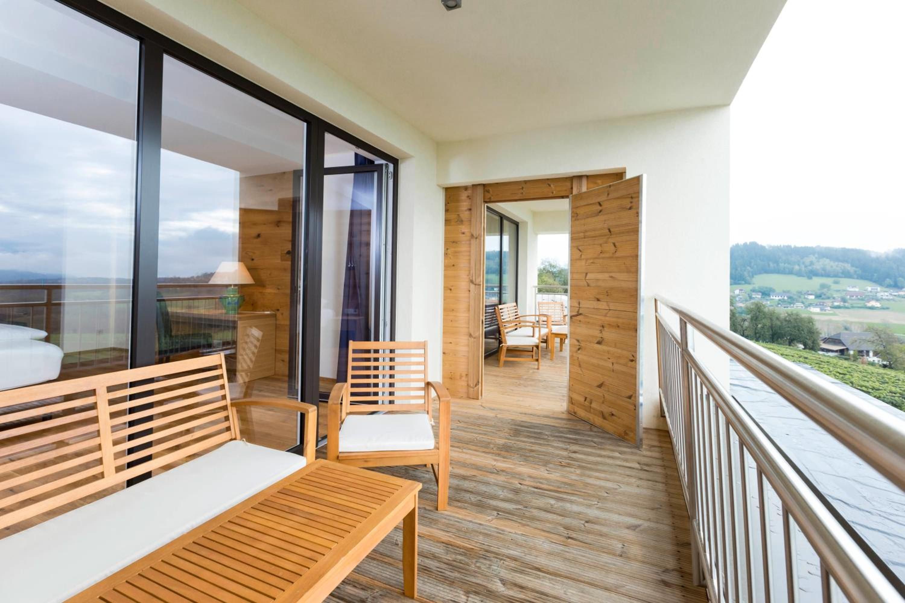 A balcony with wooden furniture, a glass door leading to an outdoor area, and a view of a landscape.
