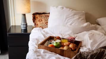 Un llit ben fet amb roba de llit blanca, una safata de fusta amb articles per a l'esmorzar, com ara croissants, una poma i un got de suc d'aranja, i una làmpada sobre una taula de nit.