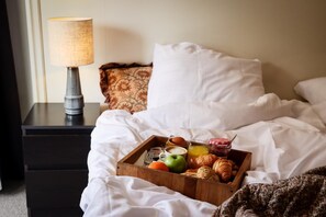 Cama bem arrumada com lençóis brancos, bandeja de madeira com itens de caf é da manhã como croissants, uma maçã e um copo de suco de laranja, e um abajur em uma mesa de cabeceira.