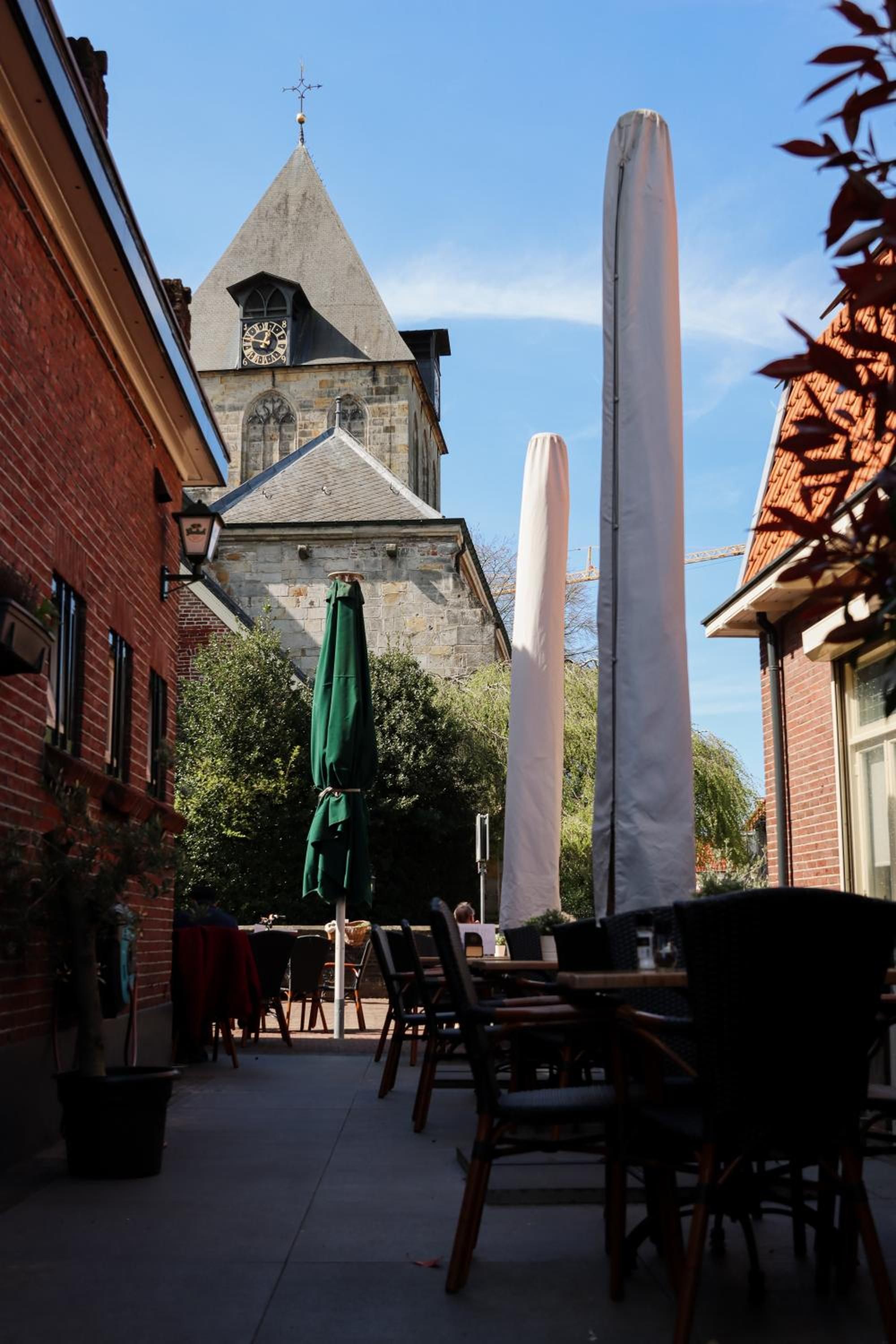 Un área de estar al aire libre con mesas y sillas, un paraguas verde y un edificio con una torre de reloj al fondo.