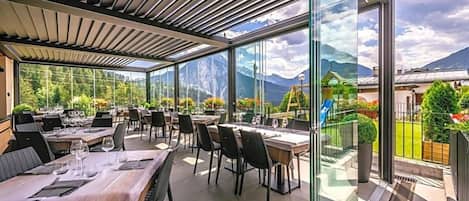 Un área de comedor al aire libre con mesas y sillas, ventanas amplias y vistas a las montañas.