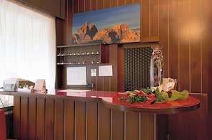 A reception desk with a wooden finish, a framed landscape painting above, and a decorative vase with flowers on the counter. - Hotel Bel Sit (Valle di Cadore)