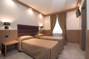 Ein Hotelzimmer mit zwei Betten, einem Nachttisch, einem an der Wand befestigten Fernseher und einem Fenster mit Vorhängen.